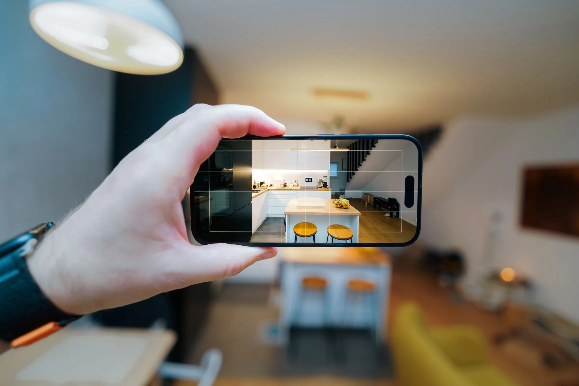 Hand hält ein Smartphone quer und fotografiert eine Küche mit Kochinsel und zwei Barhockern, das Foto ist auf dem Display zu sehen